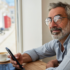 Hombre con lentes progresivas mirando por la ventana de una cafetería tras consultar el móvil, con un portátil sobre la mesa